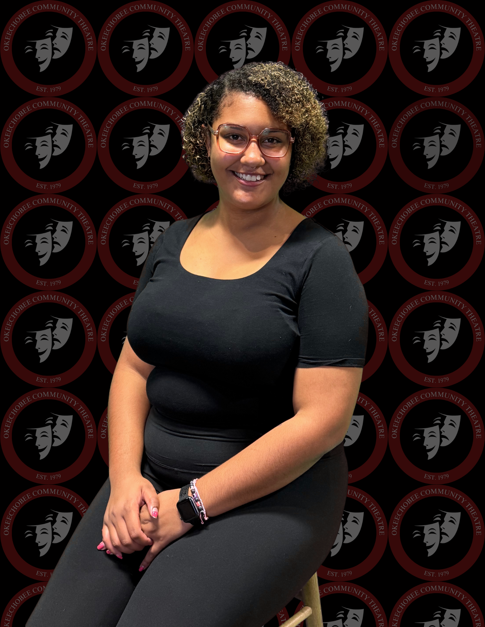 A woman smiling in front of a backdrop with the Okeechobee Community Theatre logo. She has curly blonde hair, is wearing a black polo shirt with the theater's logo, a white watch, and blue jeans.