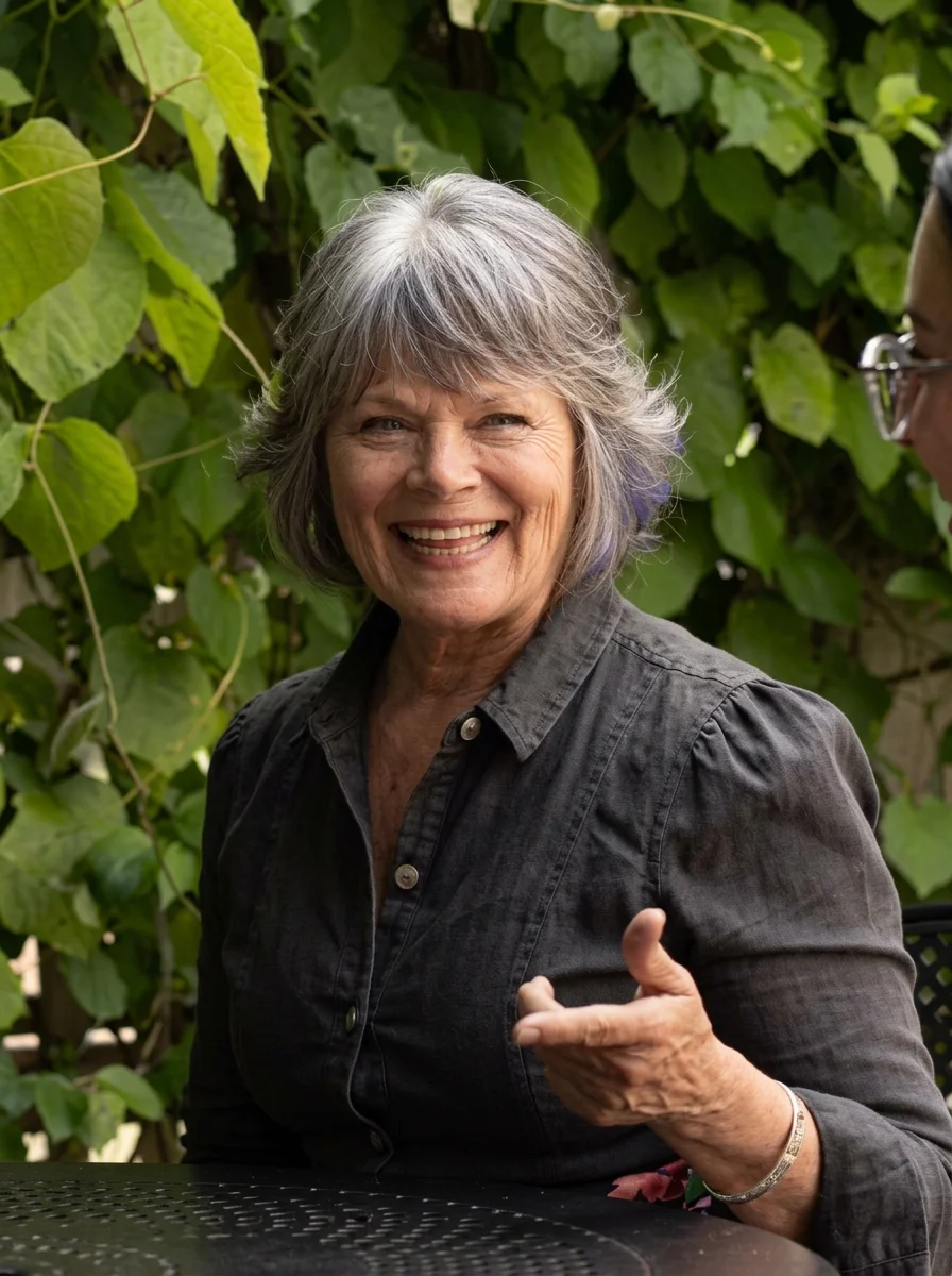 A joyful older woman with gray hair smiling and gesturing with her hand, sitting outdoors with lush green foliage in the background.