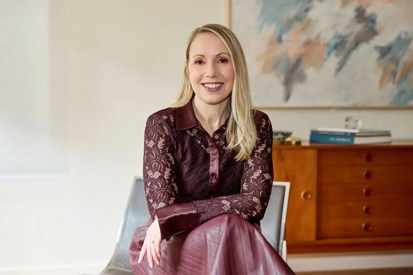 A woman with blonde hair, wearing a maroon lace top and matching skirt, sitting in a modern room with a wooden dresser and abstract painting behind her, smiling at the camera.