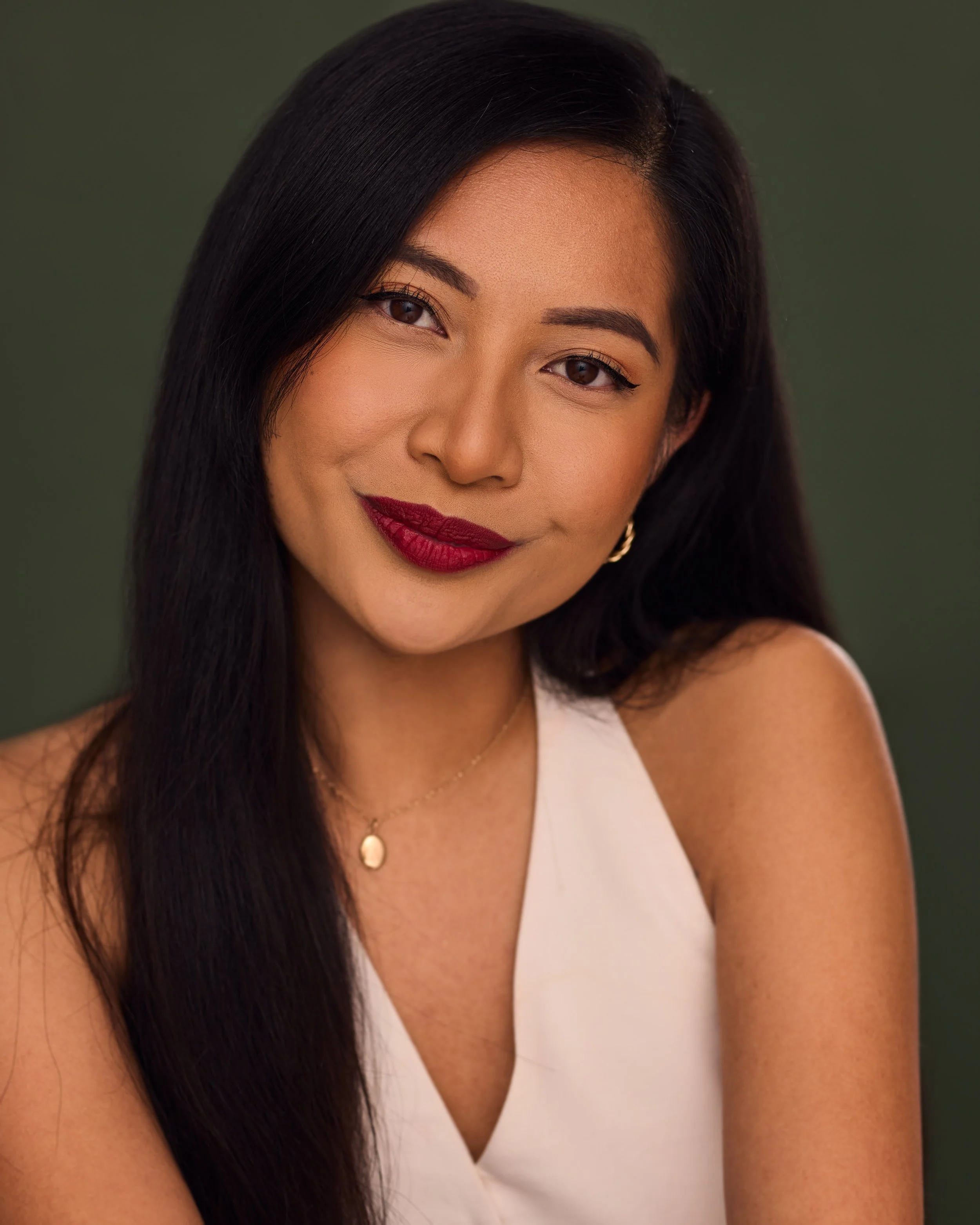 A woman with dark hair, wearing red lipstick, a white sleeveless top, and gold jewelry, smiling at the camera against a dark green background.