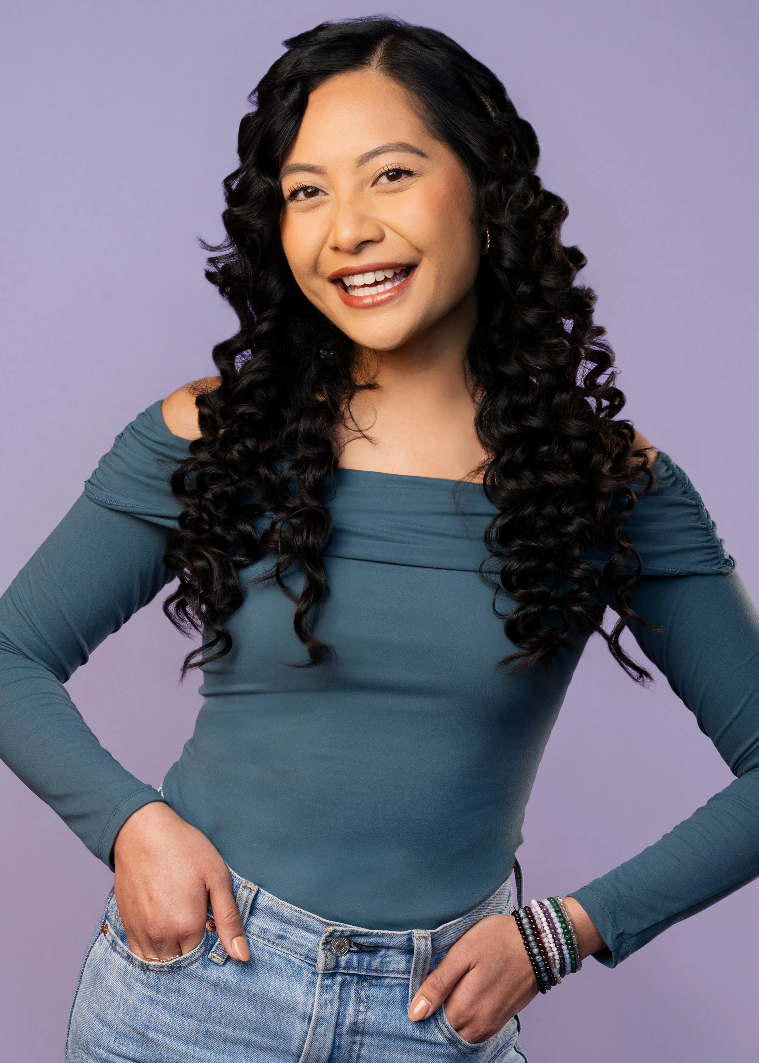 A woman with long black curly hair, smiling and winking, wearing a teal off-the-shoulder top and light blue jeans, standing against a purple background.
