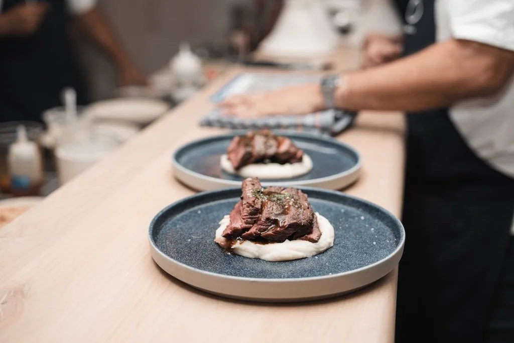 Two plates of steak on creamy white mashed potatoes being prepared by cooks in a kitchen setting.