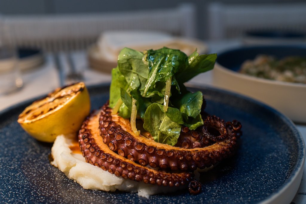 Grilled octopus on mashed potatoes with grilled lemon and greens on a blue plate.