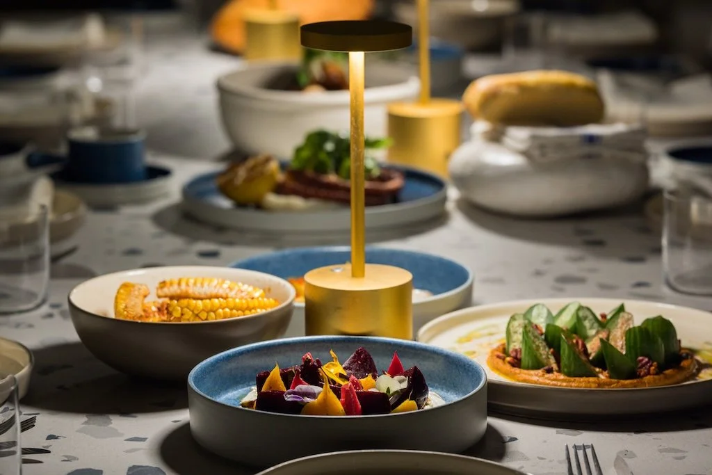 A dining table set with gourmet dishes and a modern lamp in the center. The table features plates with colorful food, including corn and vegetables, accented by elegant table settings and soft lighting.