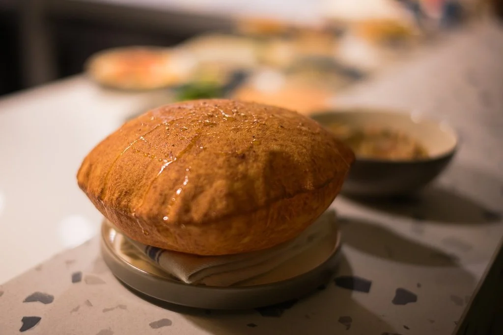A large, golden-brown, inflated round bread on a plate with a bowl of food in the background.