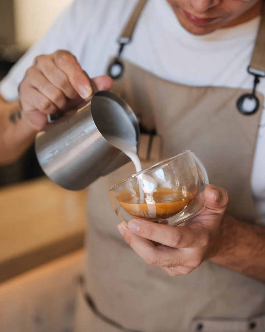 Man in beige apron pouring milk into espresso in a glass cup.
