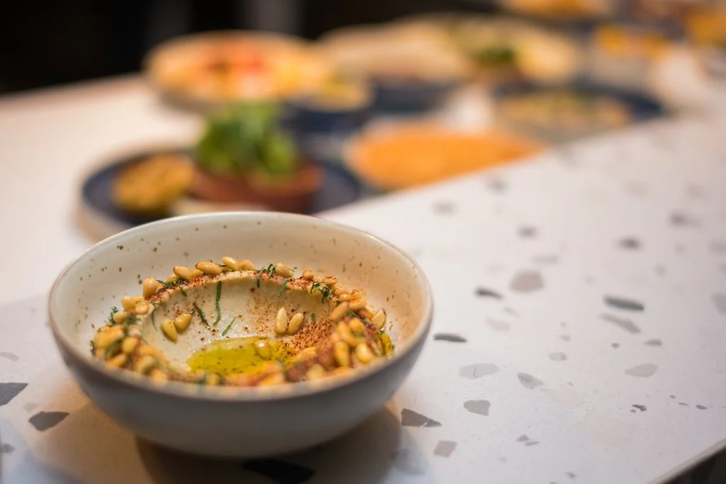 Bowl of hummus topped with pine nuts and herbs on a countertop, with blurred dishes in the background.