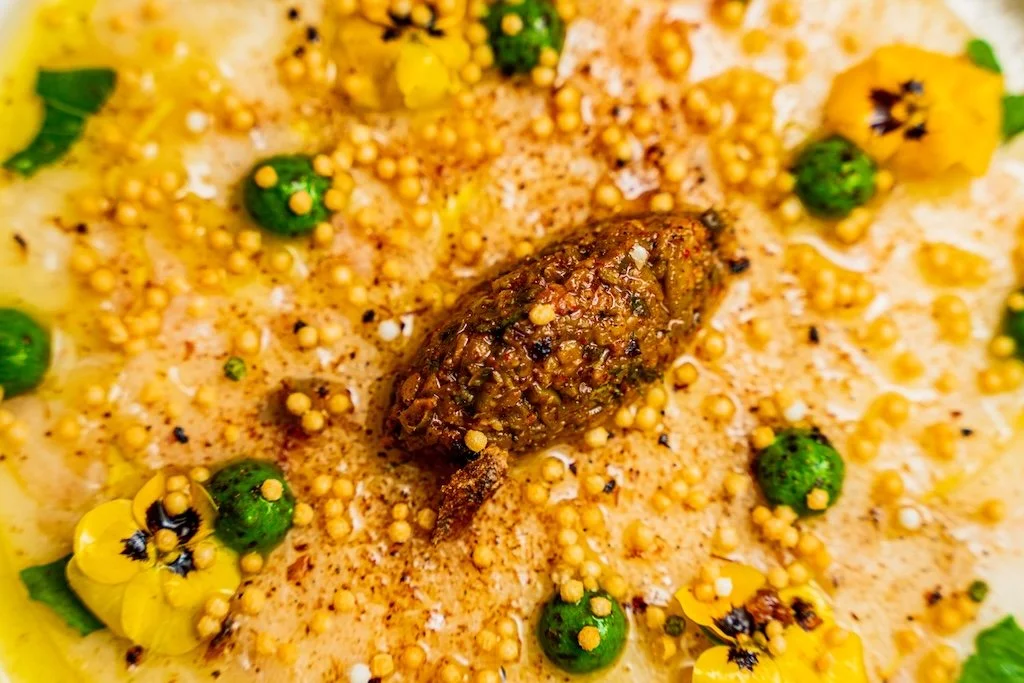 Gourmet dish with edible flowers, spices, and a brown central piece surrounded by green drops and mustard seeds.