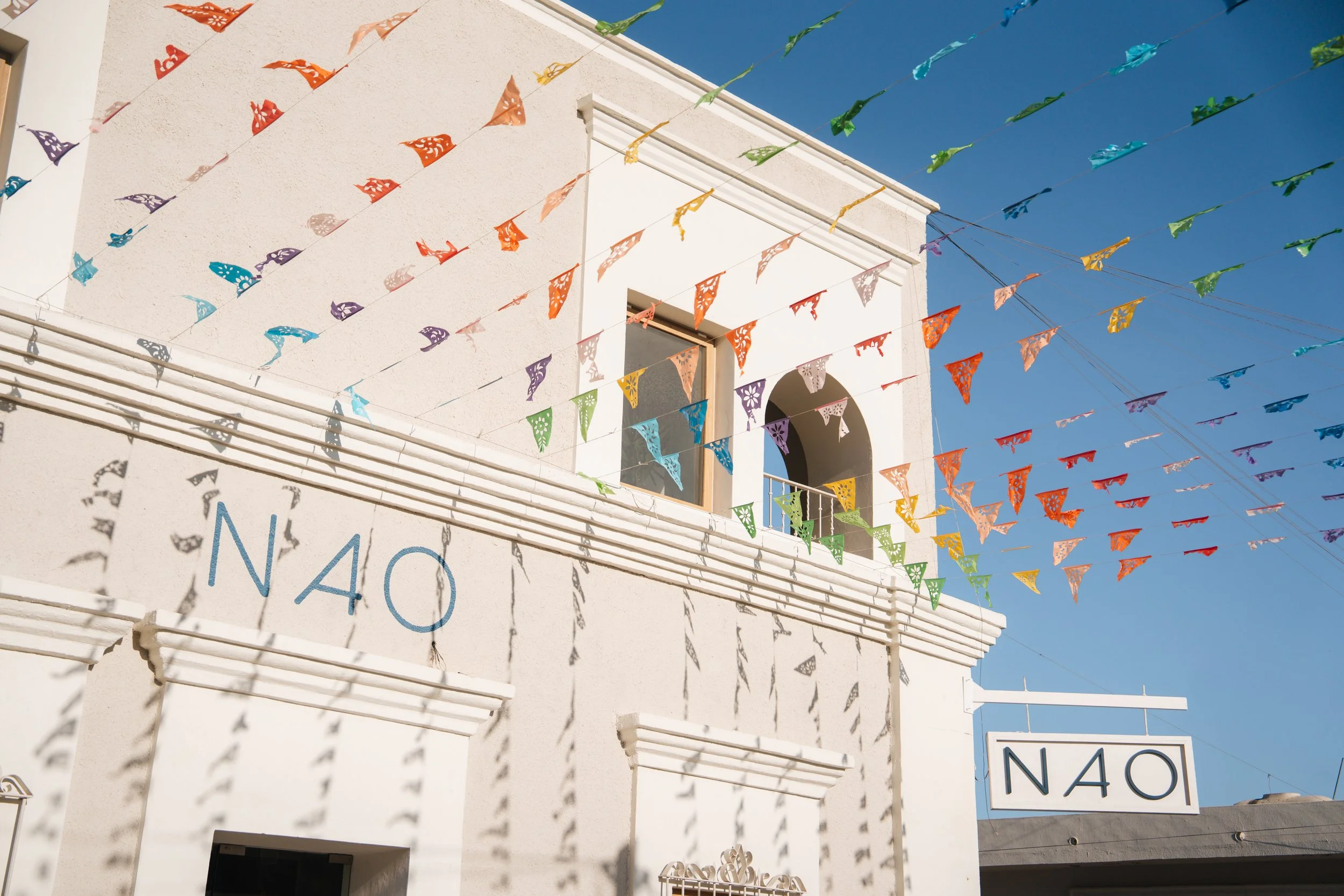 Colorful papel picado banners hanging above a white building with 'NAO' signage against a clear blue sky.
