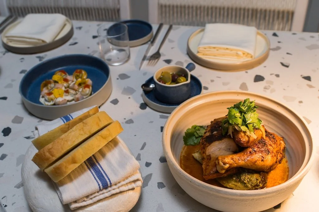 Dining table with roasted chicken, plate of bread, olives, and a small salad.