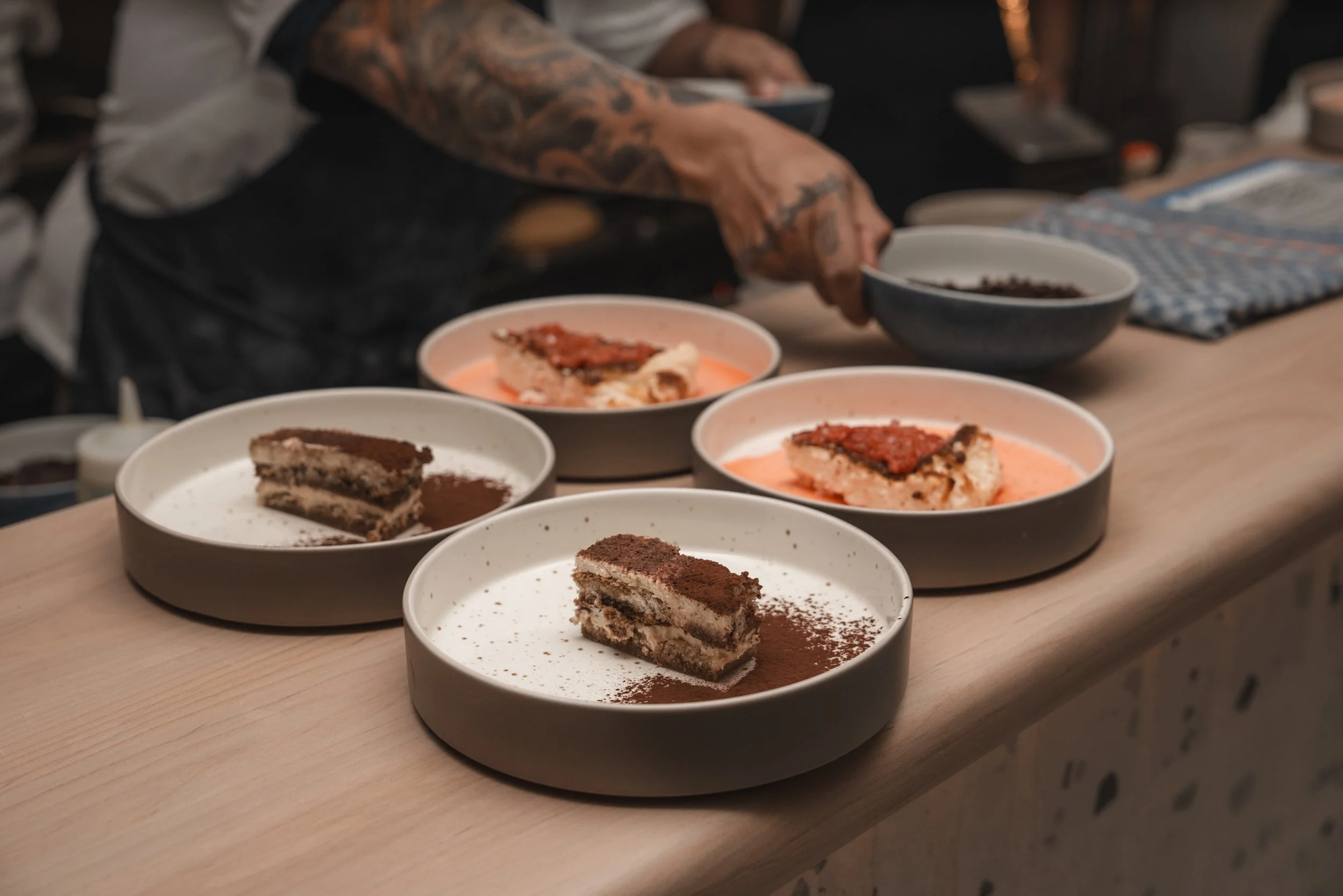 Plated desserts with tiramisu and another dessert, with a person's tattooed arm reaching over.