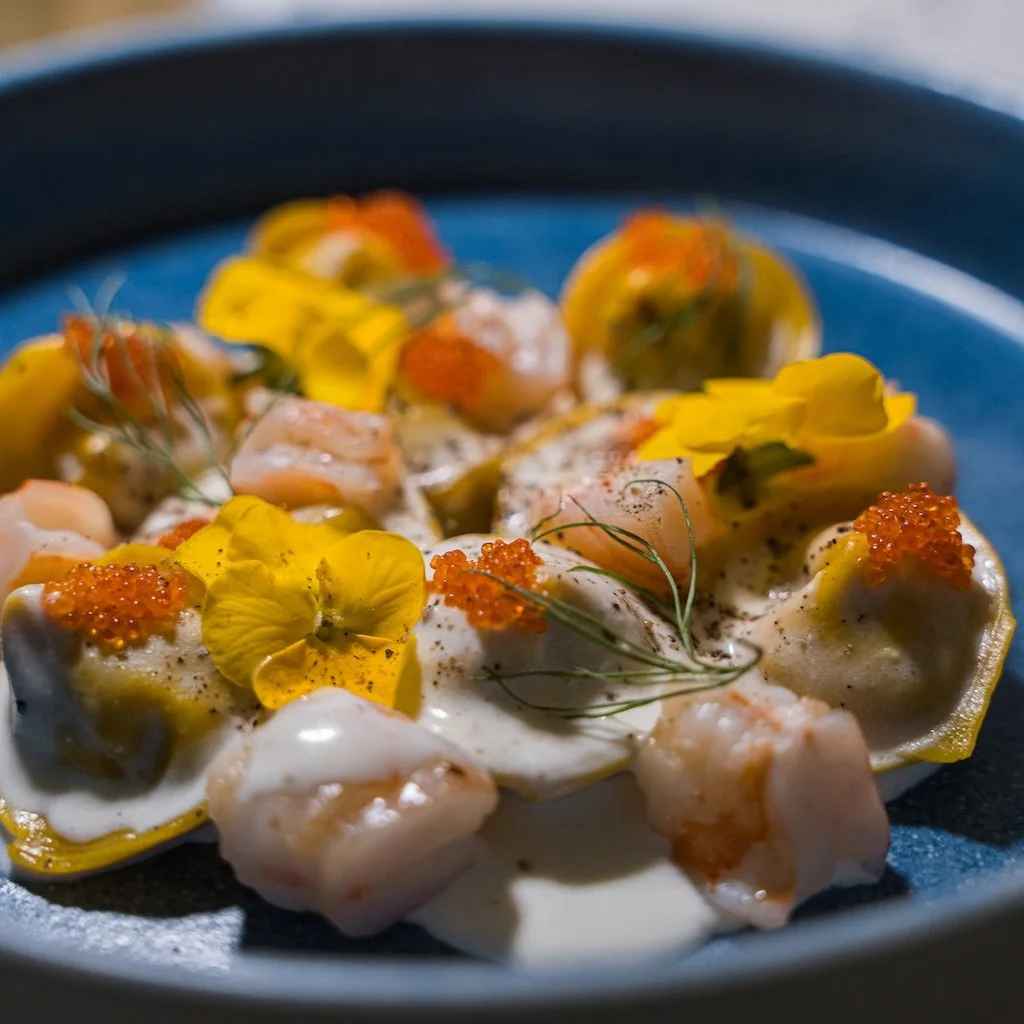 Gourmet dish with ravioli, shrimp, yellow edible flowers, orange fish roe, and cream sauce on a blue plate.