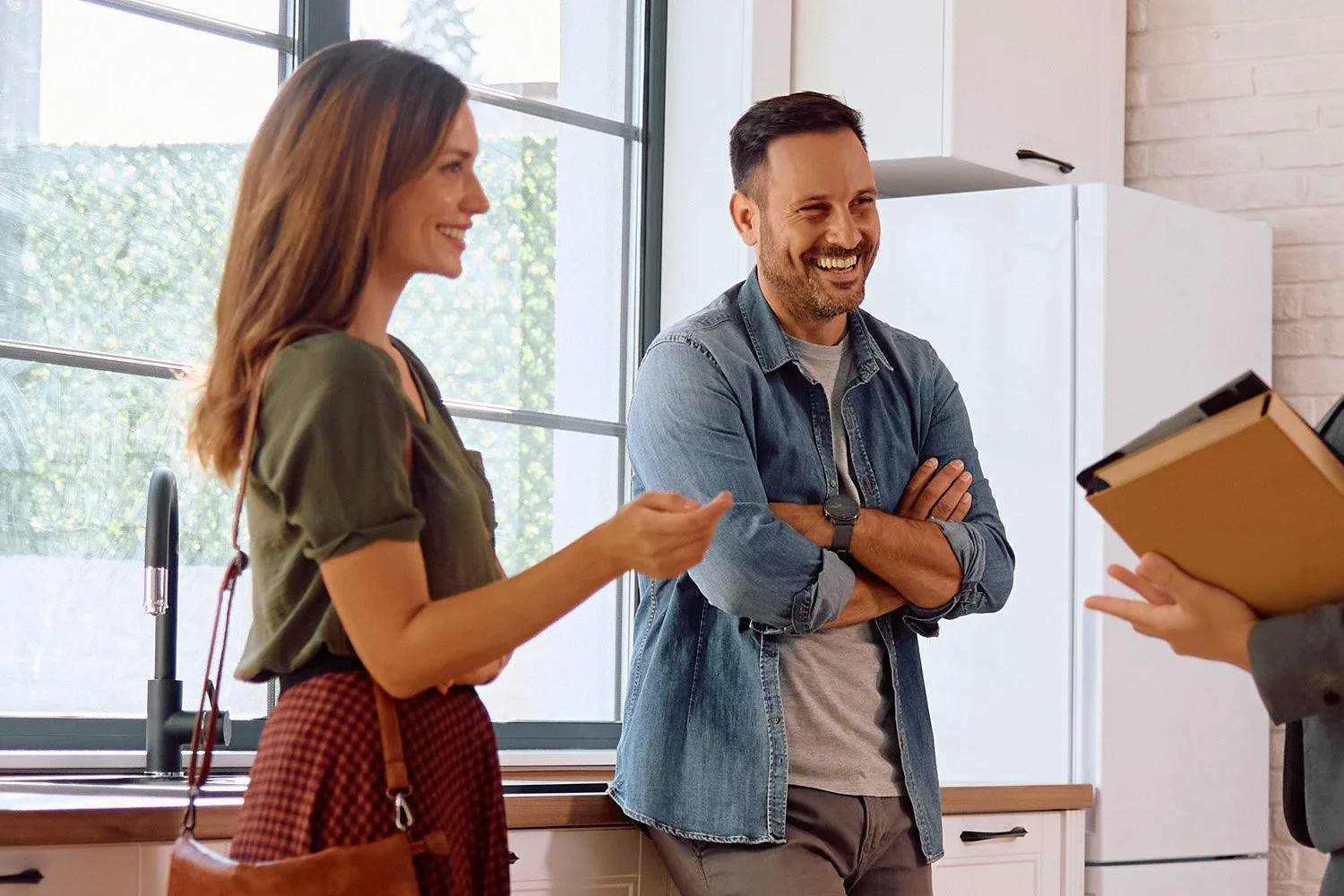 Two smiling people, a woman and a man, are having a conversation in a well-lit kitchen, while a third person holds a clipboard nearby.