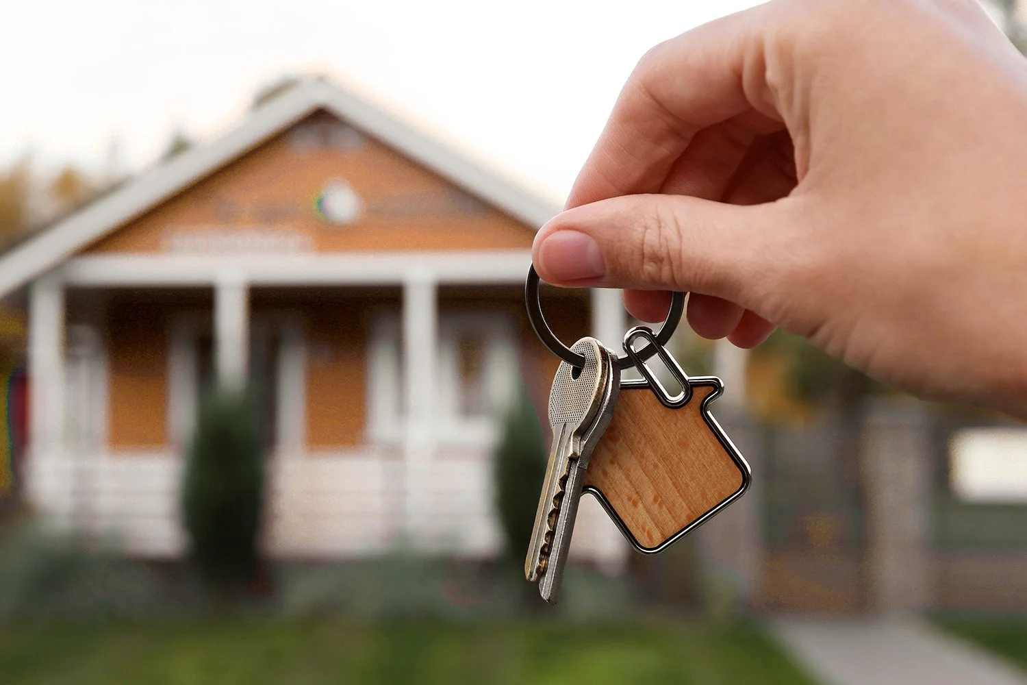Person holding a keychain with house-shaped key and house-shaped key fob in front of a house.