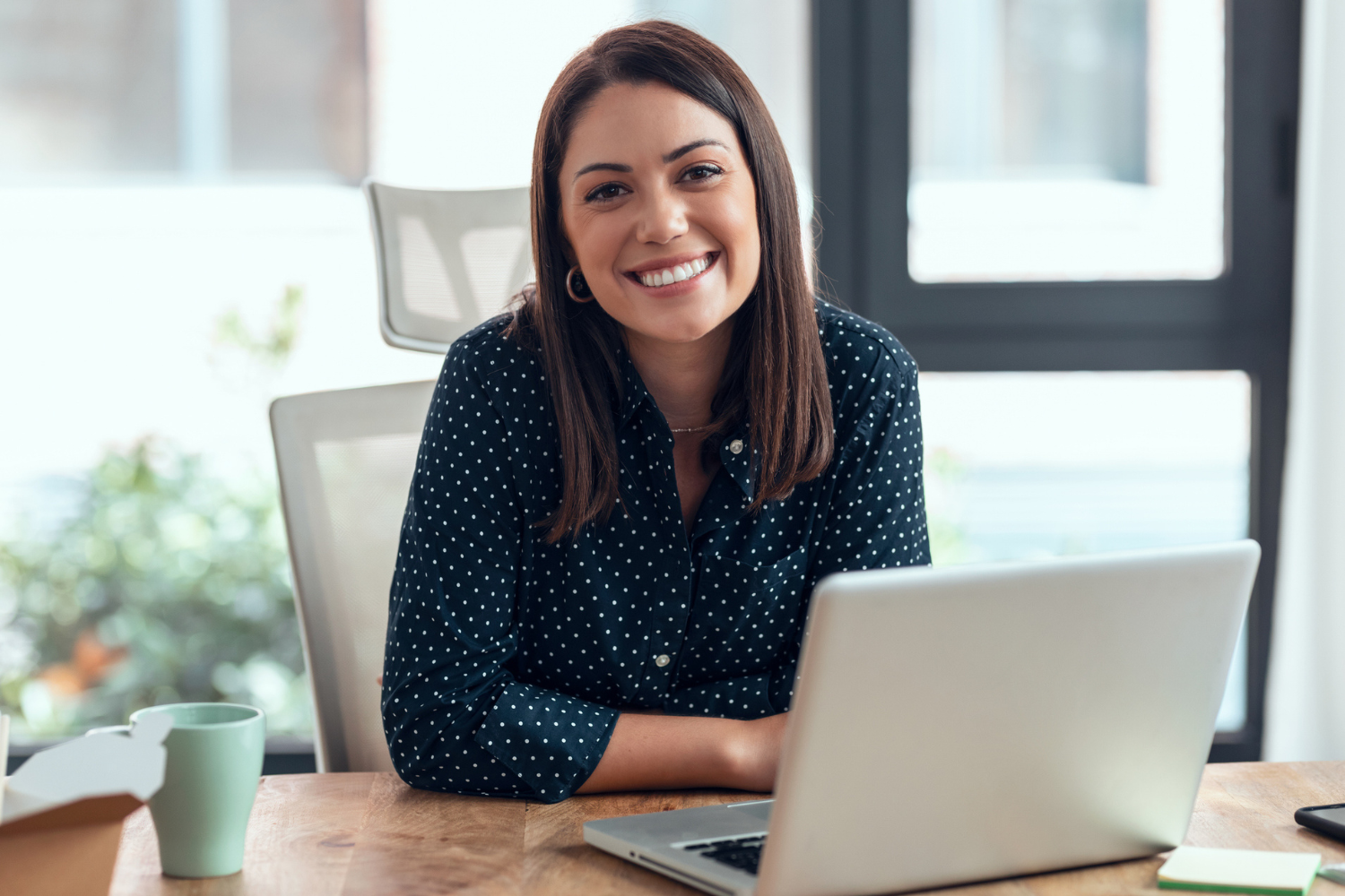 A young woman with shoulder-length brown hair, wearing a navy blue polka dot shirt, sitting at a wooden desk with a laptop, smiling at the camera in a bright, modern office.
