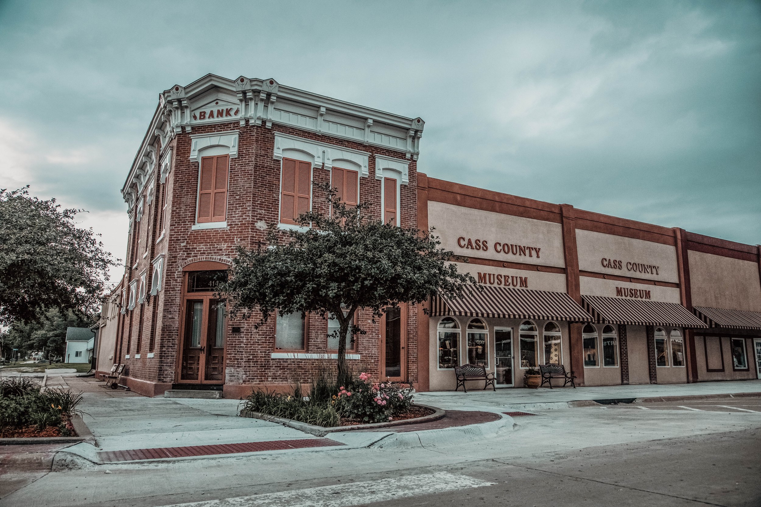 Exterior of Cass County Museum and Bank building with brick and beige walls, striped awnings, trees, and benches in front.