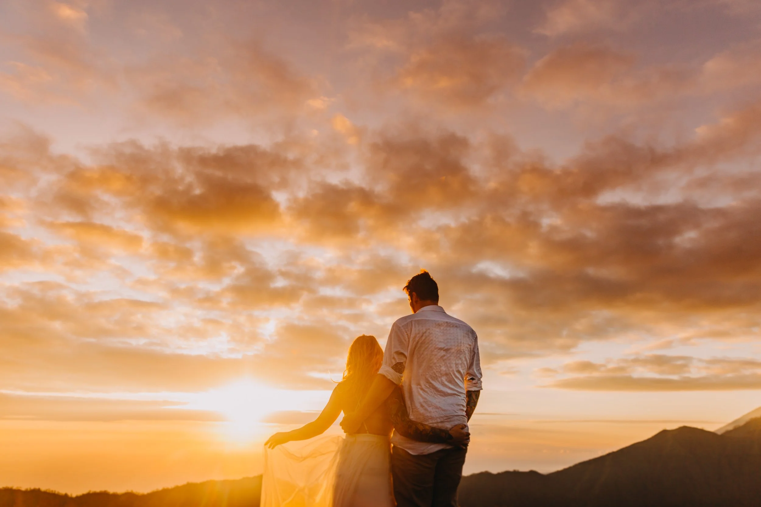 A couple stands close together at sunset, facing the horizon with a scenic landscape and mountains in the background, while the sky is filled with orange and pink clouds.