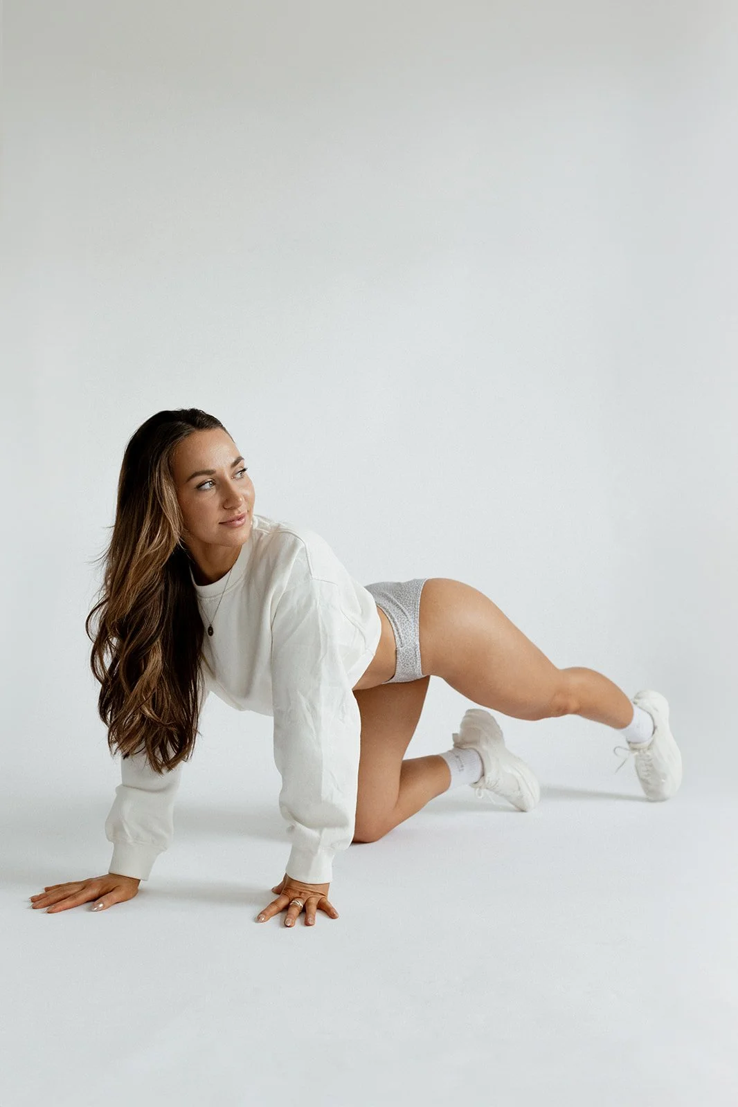 A woman in a white sweatshirt, gray shorts, and white sneakers on all fours, looking to the side against a plain white background.