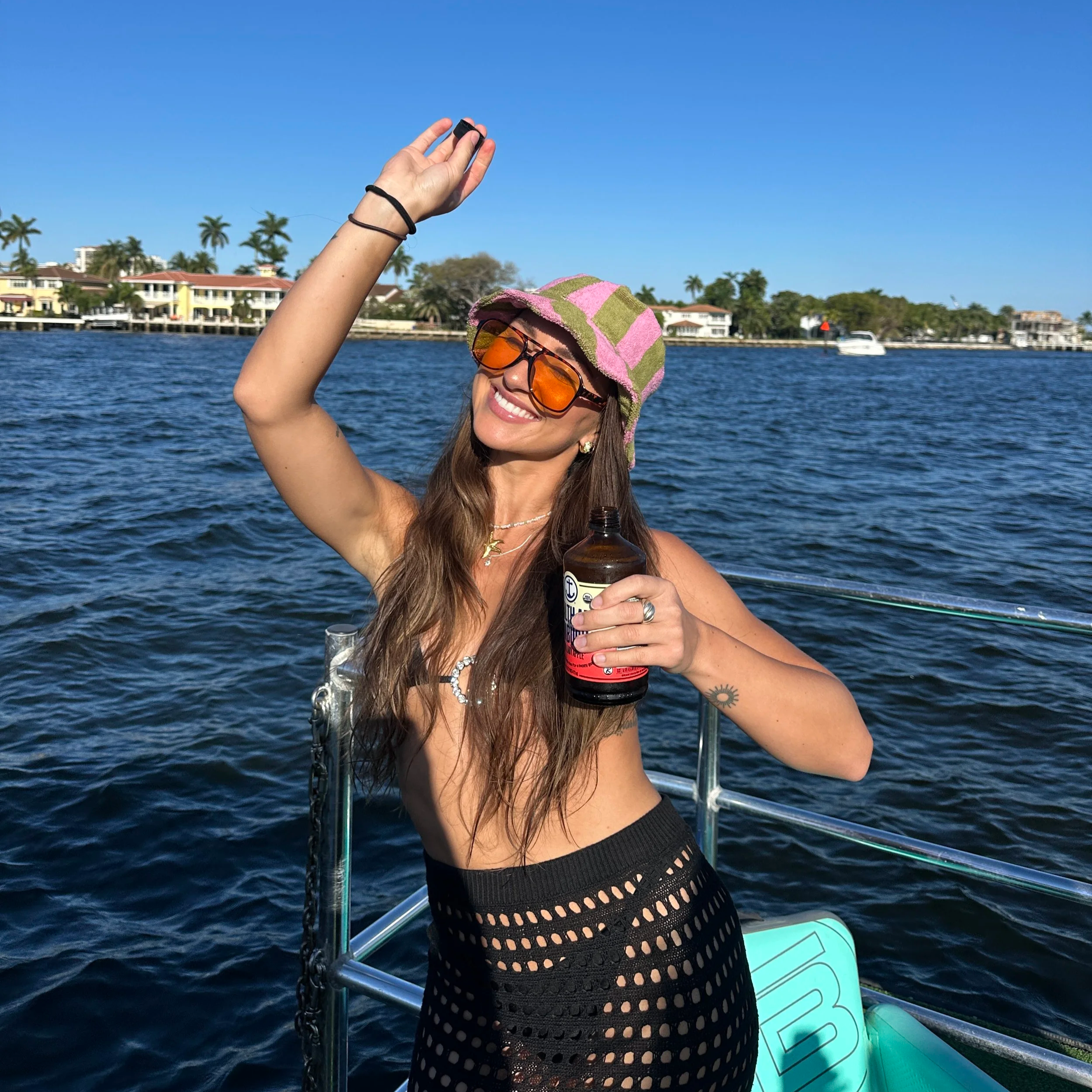 A woman on a boat, smiling and holding a drink, wearing sunglasses, a colorful hat, and a black crochet skirt, with water and houses in the background.