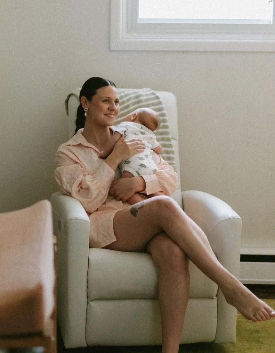A woman with dark hair, wearing a pink shirt and shorts, sitting in a white armchair, holding a sleeping baby wrapped in a blanket, in a room with a window and a heater.
