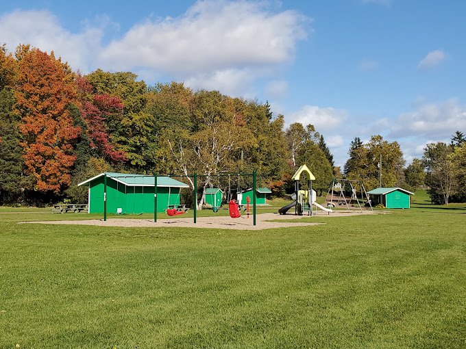 Playground with green structures including swings, slides, and a picnic area surrounded by autumn trees under a blue sky.