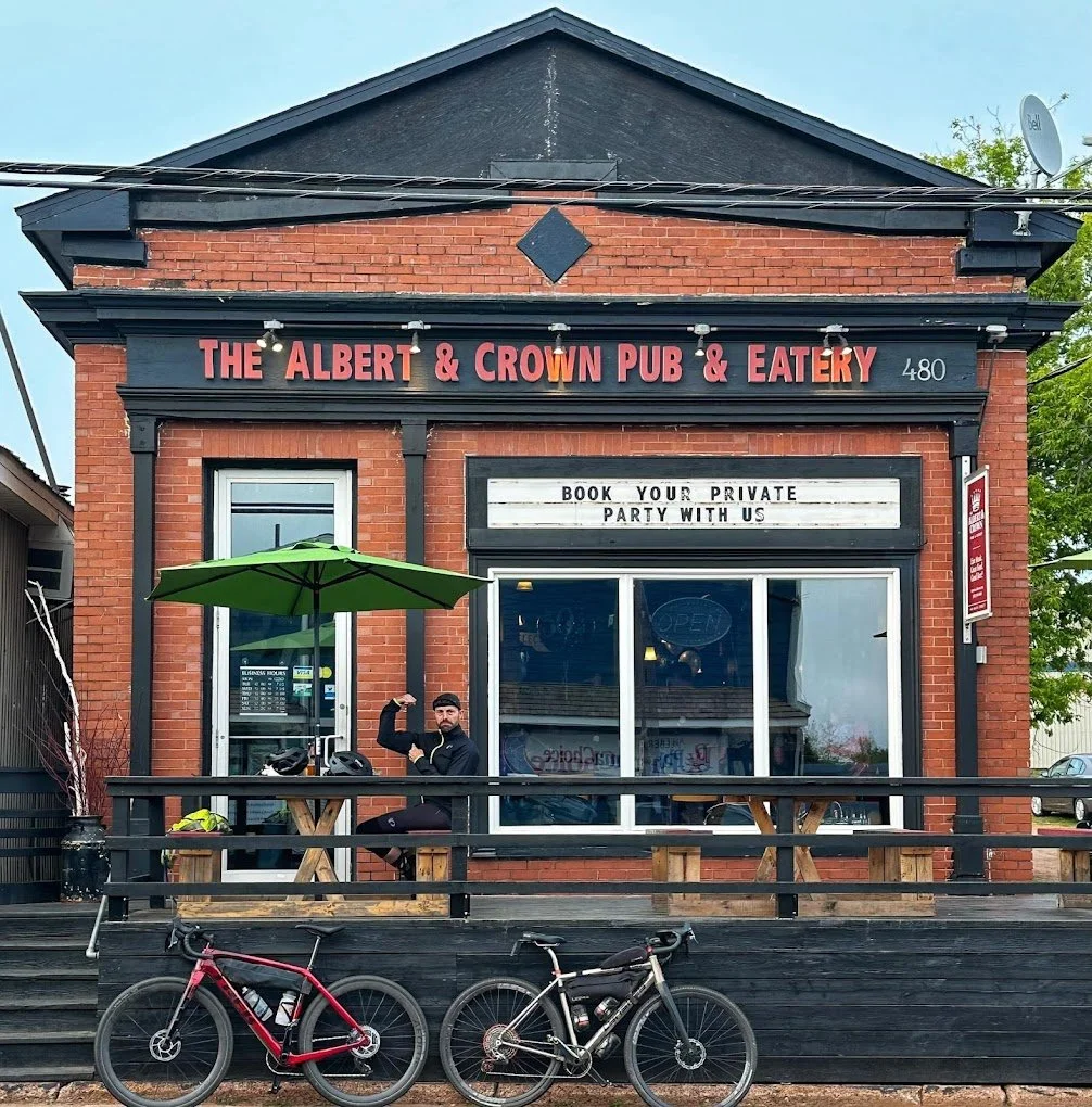 Exterior of The Albert & Crown Pub & Eatery with a brick facade. There's a person sitting at a table on the patio under a green umbrella. Two bicycles are parked in front. Sign reads 'BOOK YOUR PRIVATE PARTY WITH US.'