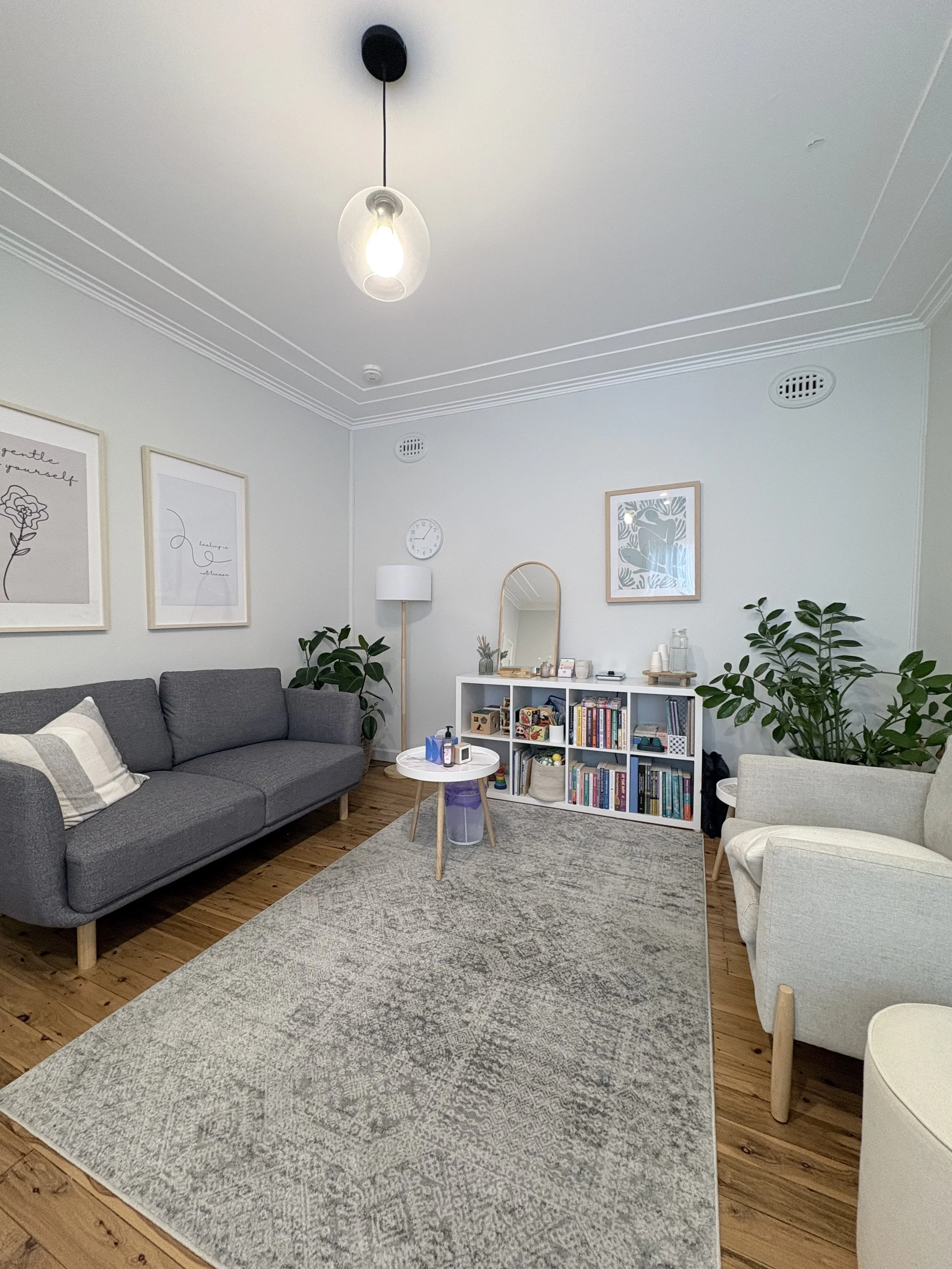 A cozy living room with a grey sofa, cream armchair, a small round white and wood coffee table, a white bookshelf filled with books, and large green potted plants. The room has light-colored walls, wooden flooring, and framed art on the walls. There is a ladder-style mirror and a white standing lamp, with a ceiling light.