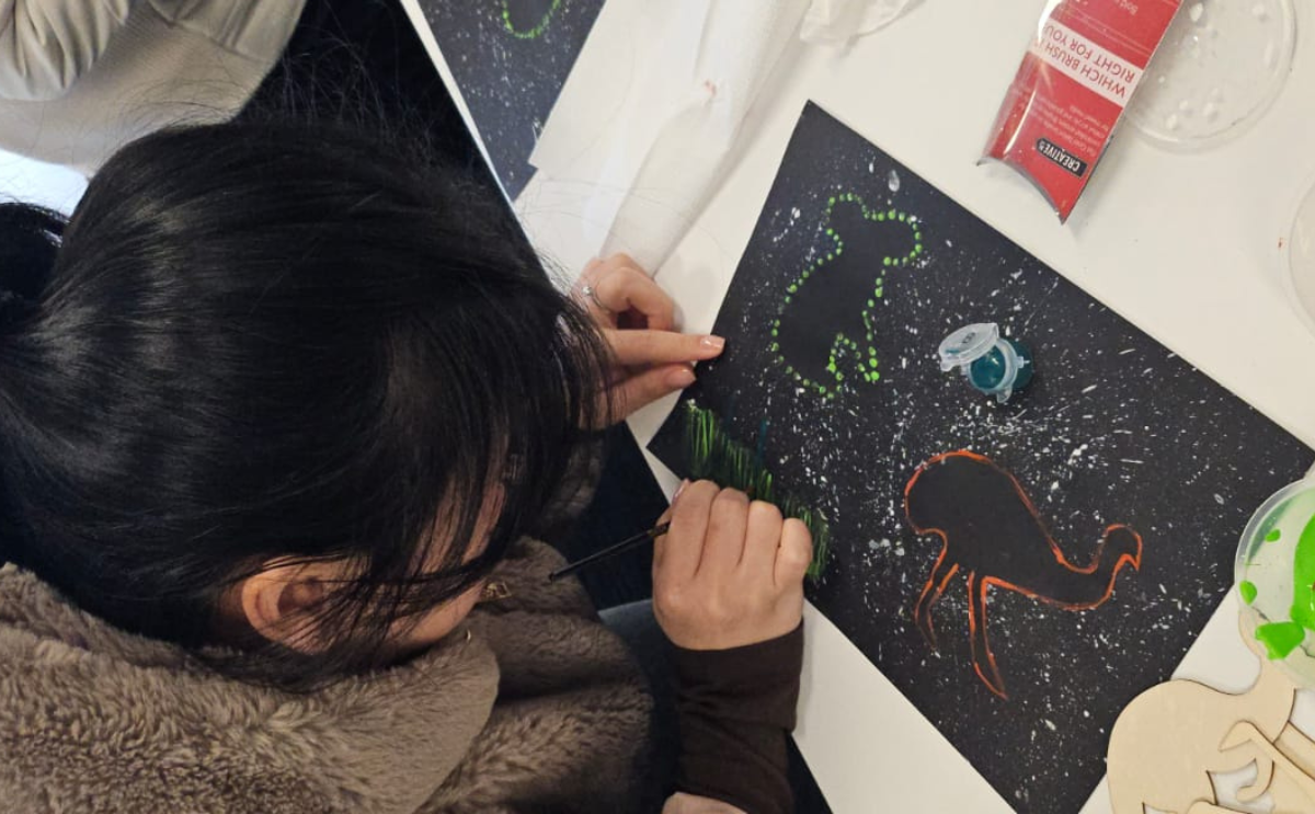 an AMEP participant with dark hair carefully painting green grass on black cardstock featuring First Nations-inspired dot art and stencils.