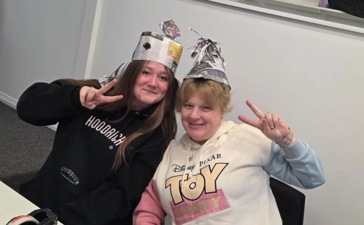 Two women wearing handmade newspaper hats smiling and making peace signs during an activity