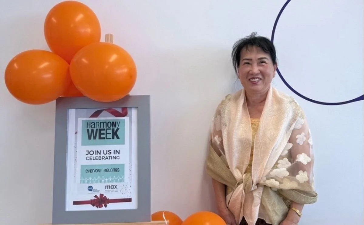 Leng, a SEE Program participant, smiling next to a Harmony Week display sign decorated with orange balloons.
