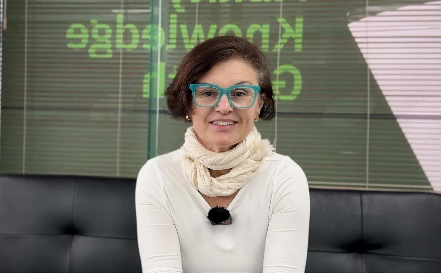 Ipek wearing teal glasses and a white top, smiling in front of a glass window displaying AKG branding.