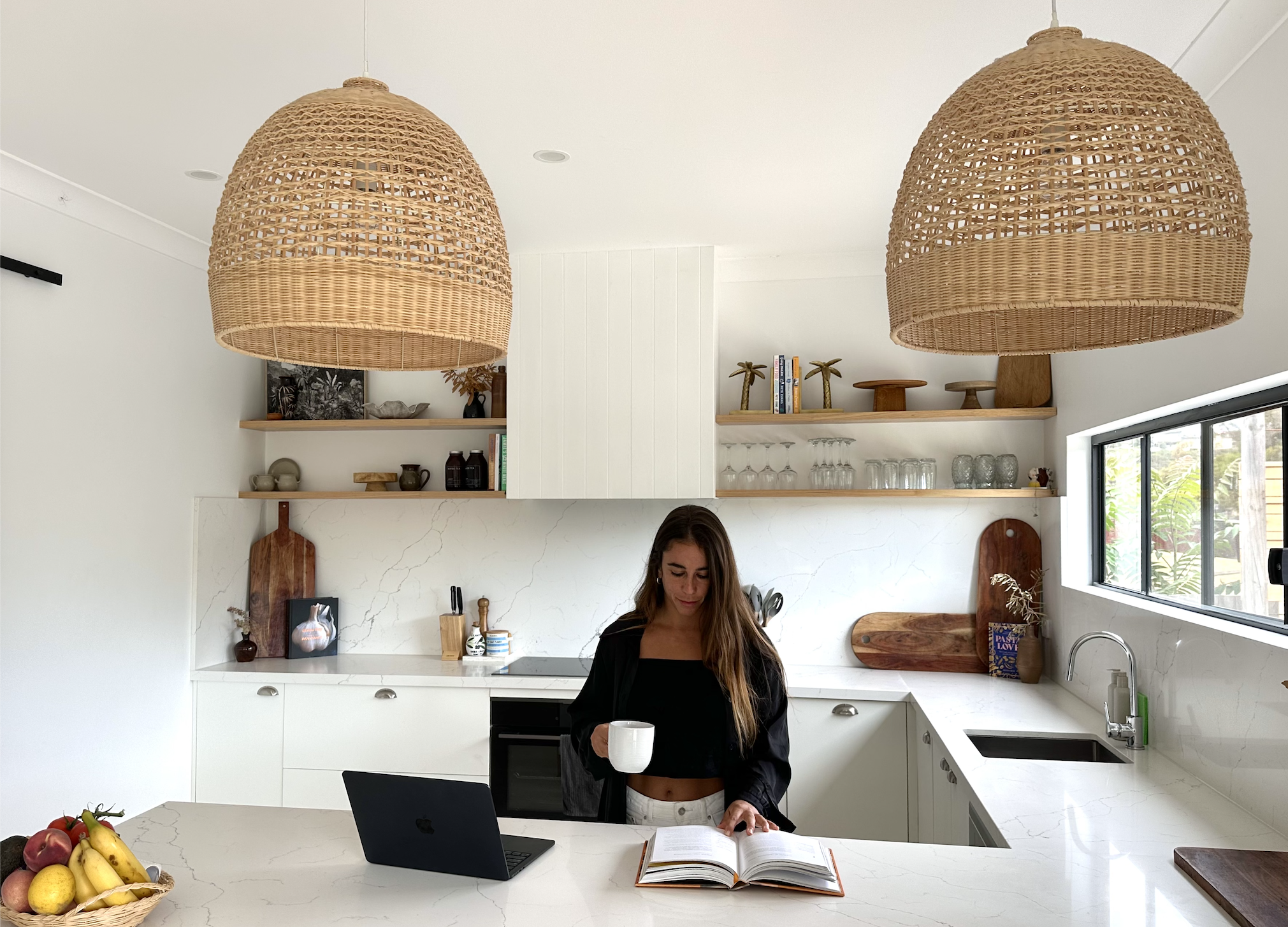 A woman in a black top and white pants standing in a bright modern kitchen, reading an open book on a white marble countertop while holding a white mug. There is a laptop in front of her, and the kitchen has wooden shelves with glassware and decorative items, a window to the right, and two large wicker pendant lights overhead.