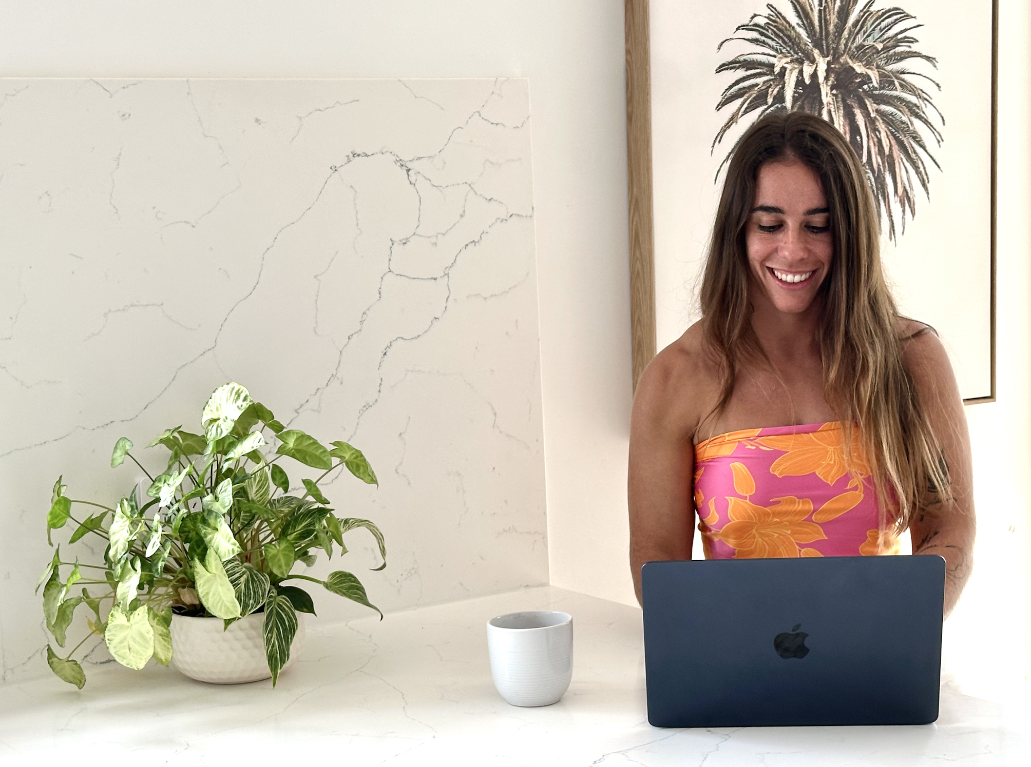 A woman with long brown hair smiling while looking at a black MacBook on a white surface, with a green potted plant and a white mug nearby, in a room with white and beige walls decorated with a palm tree artwork.