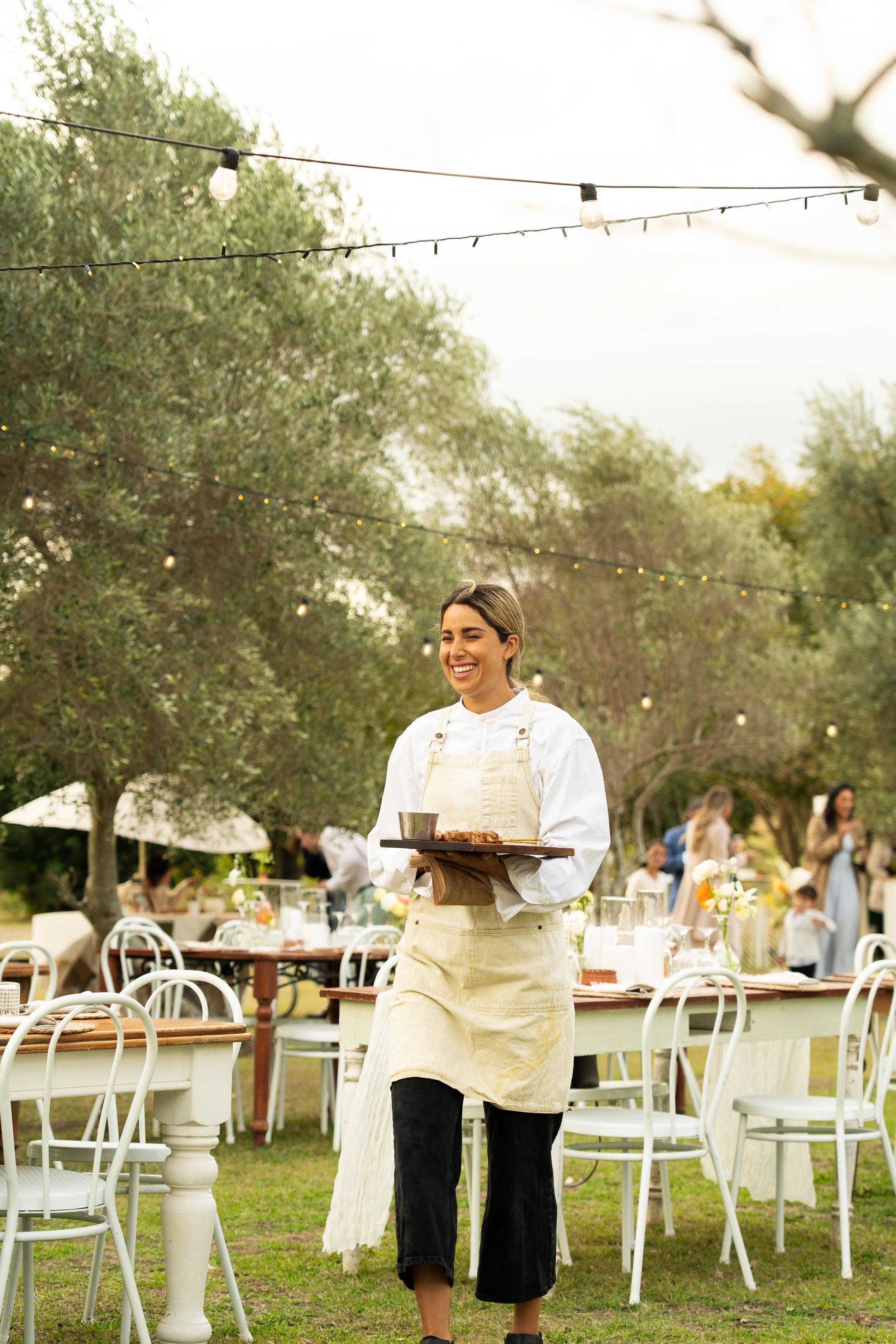 A smiling woman in a white shirt and beige apron carrying a tray with food at an outdoor gathering with decorated tables, string lights, and trees in the background.