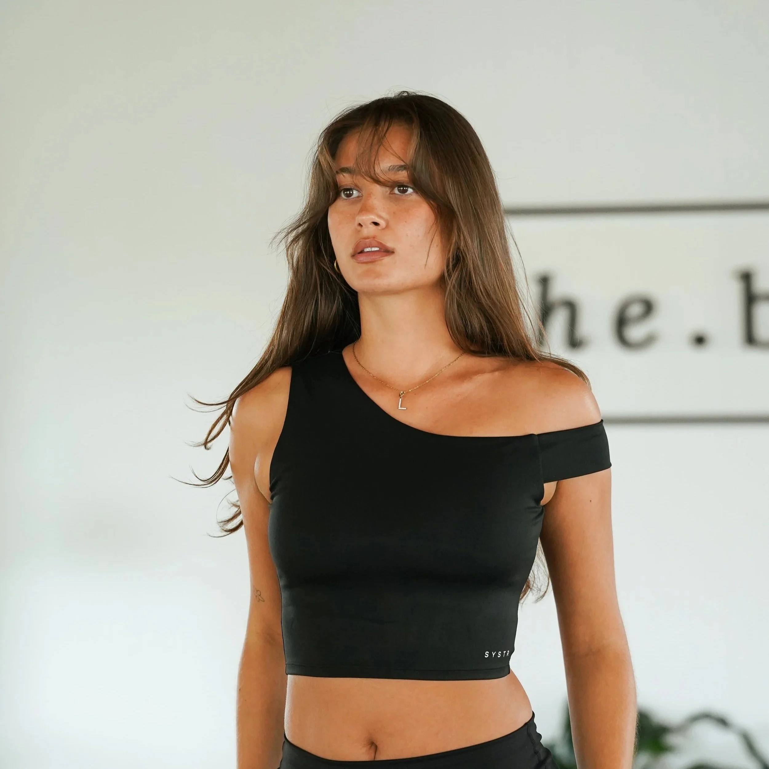 A young woman with long brown hair and tan skin, wearing a black one-shoulder crop top and gold necklace, standing indoors with a neutral expression.