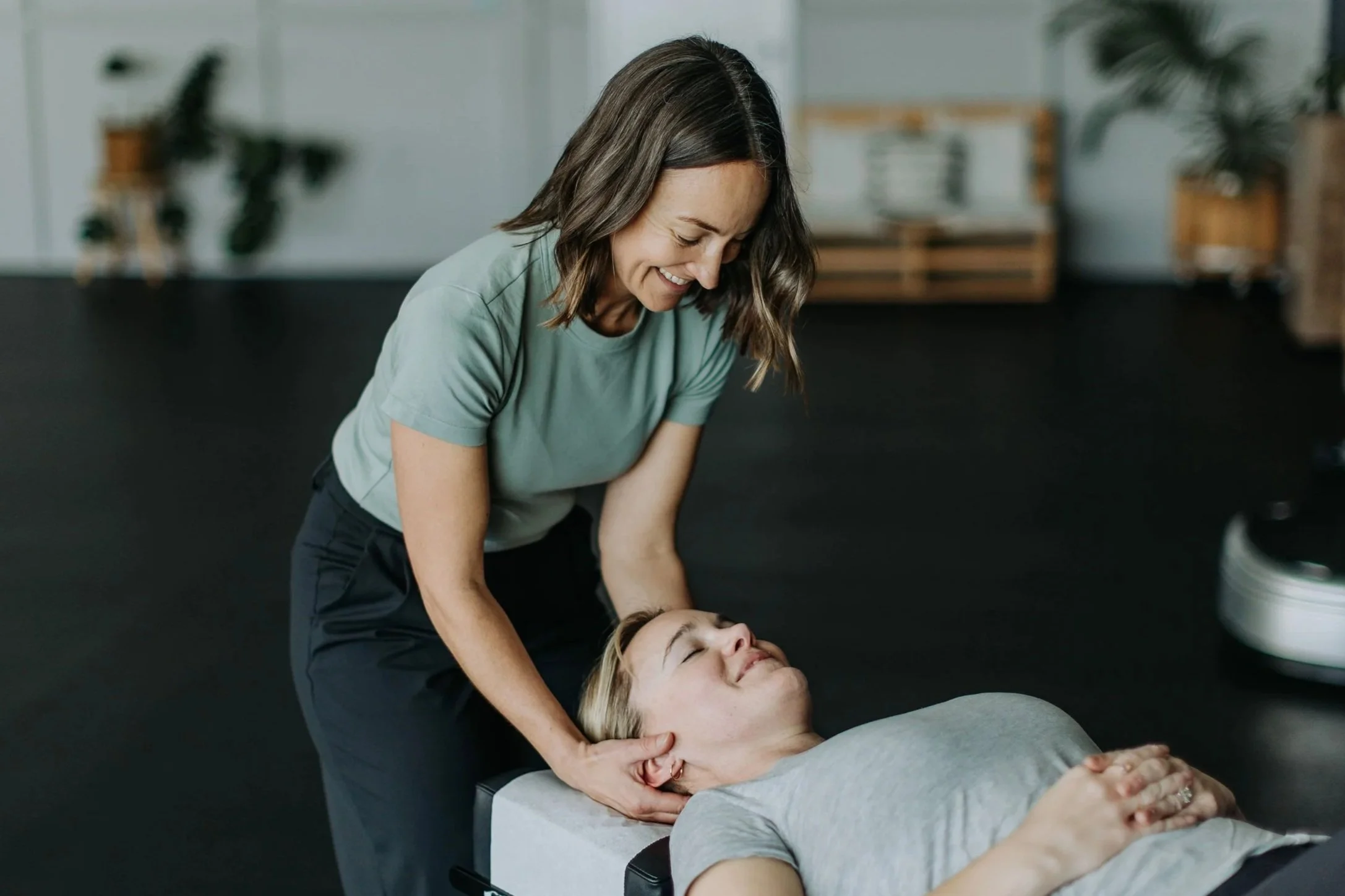A woman giving a neck massage to another woman lying on a massage table in a room with plants and wooden furniture.