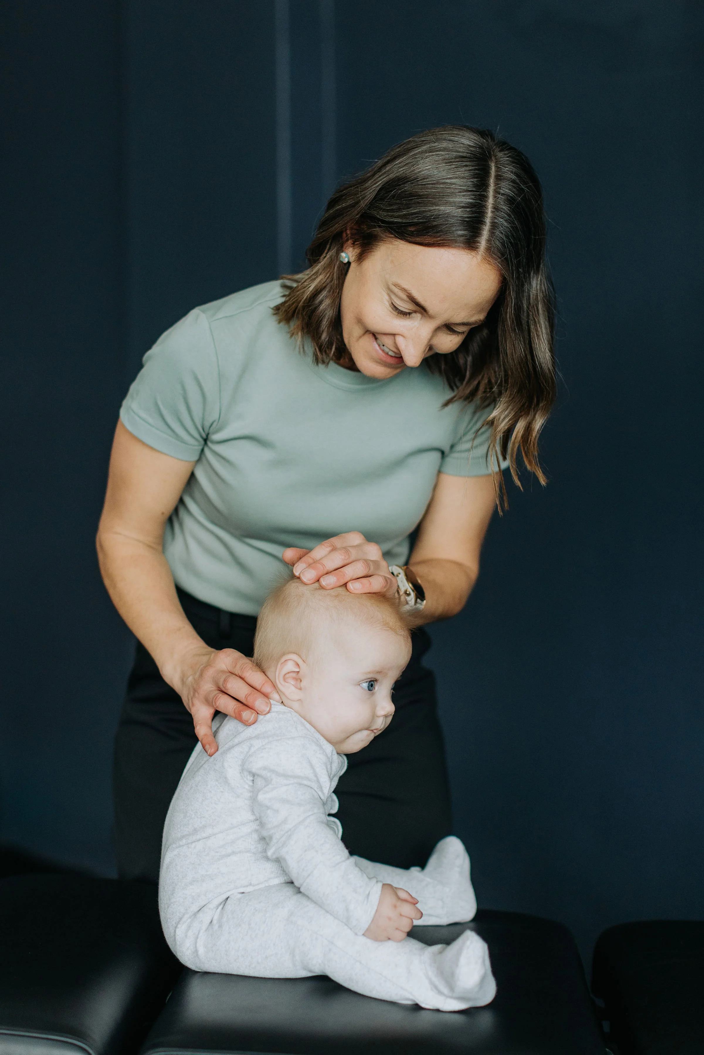 A woman with shoulder-length brown hair, wearing a light green shirt and black pants, is gently holding the head of a young child with blond hair and blue eyes. The woman is smiling and leaning over a black padded surface, while the child, dressed in