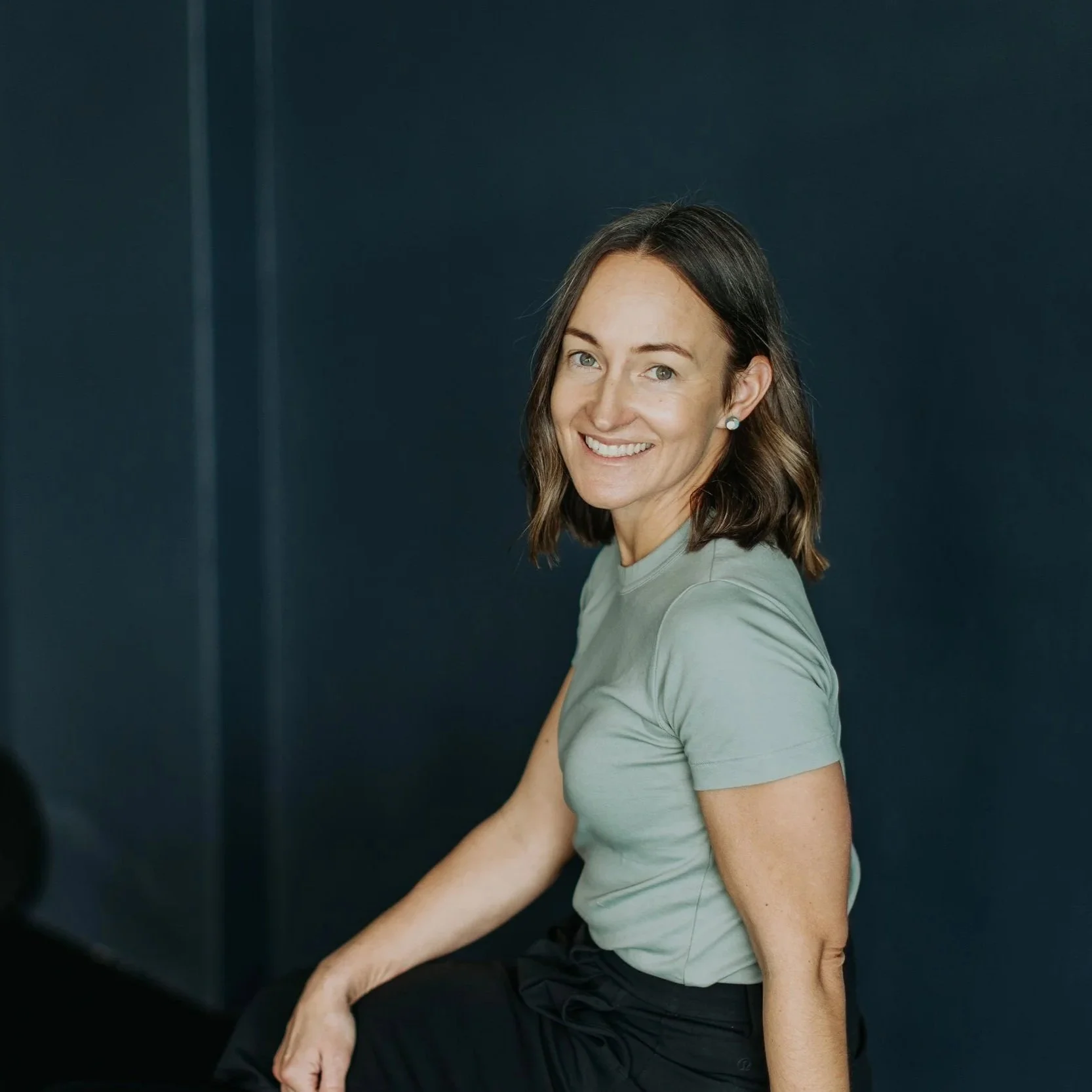 A woman with shoulder-length brown hair and light skin, smiling and looking at the camera, sitting against a dark blue background.