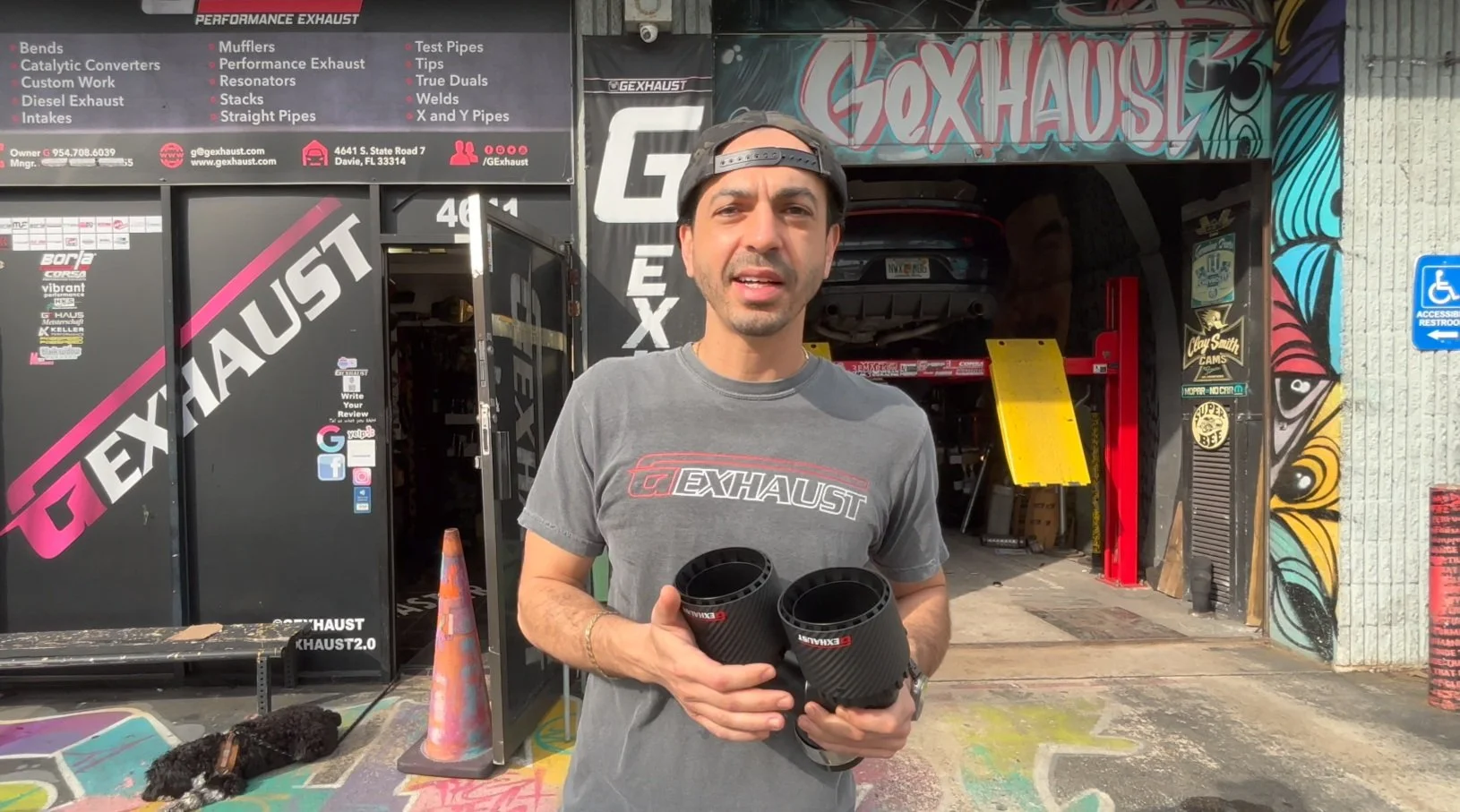 Man standing outside GEXHAUST shop holding two black exhaust tips, wearing a gray GEXHAUST t-shirt and a backward cap, with colorful graffiti on the wall behind him.