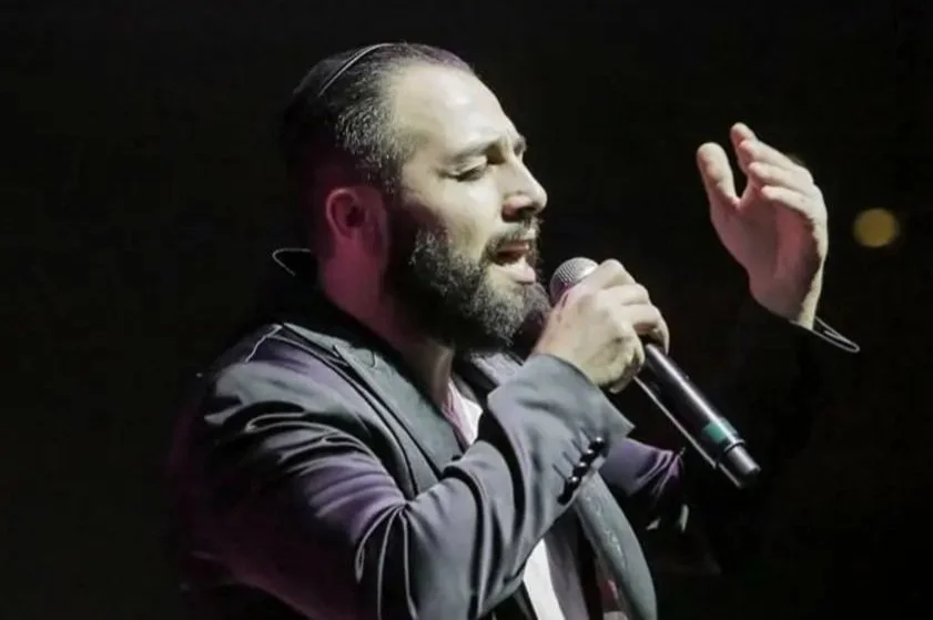 A man with a beard and slicked-back hair singing into a microphone with his eyes closed, wearing a dark jacket, with a dark background.