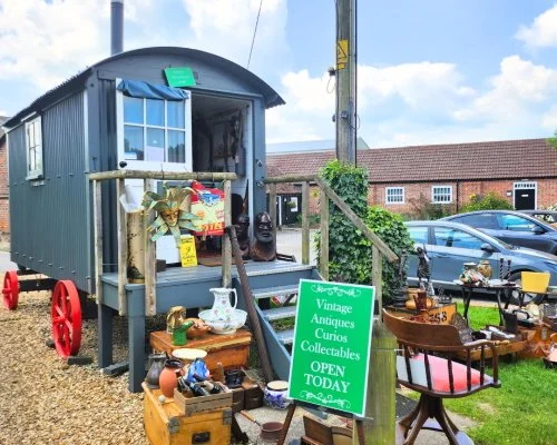 A quaint vintage shop display outside a small wagon with red wheels, featuring various antique items such as masks, pottery, and furniture. A green sign reads "Vintage Antiques Curios Collectables OPEN TODAY."