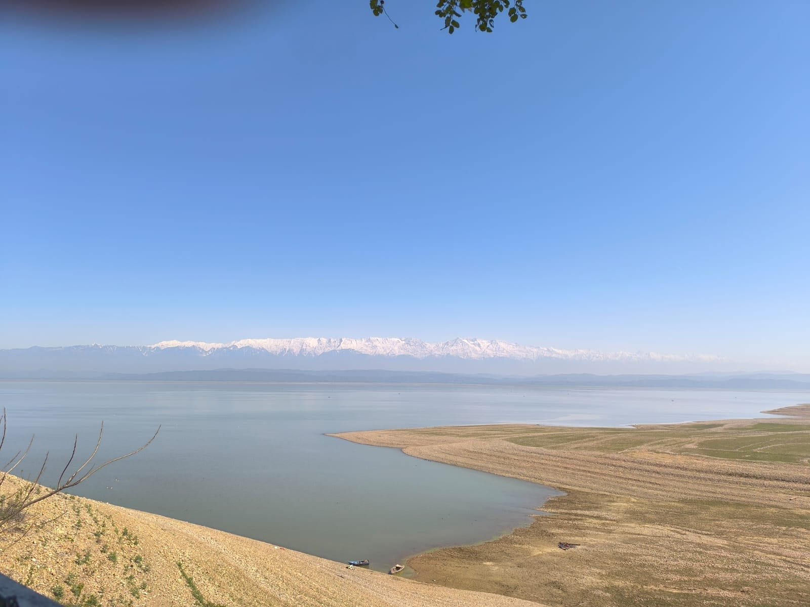 Landscape with snow-capped mountains, a large lake, and dry, grassy shoreline under a clear blue sky.