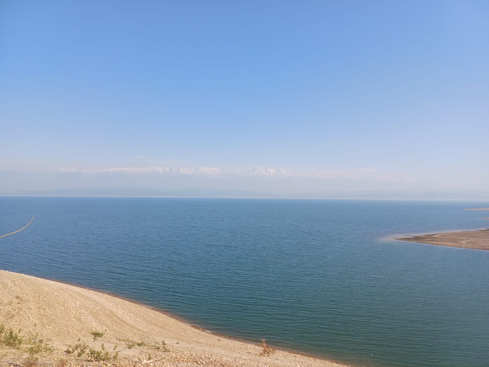 View of a large blue lake with a gravel shoreline, clear sky, and distant mountains on the horizon.
