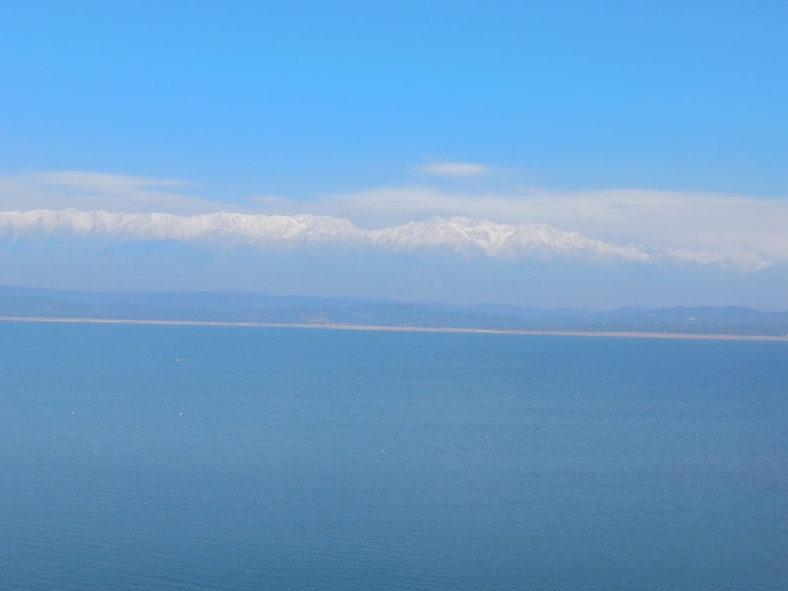 A serene lake with clear blue water in the foreground and a range of snow-capped mountains in the background under a clear blue sky.