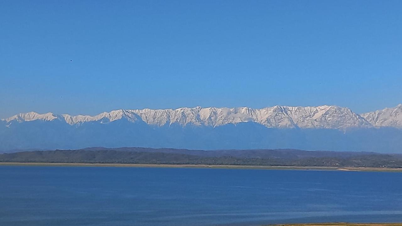 Snow-covered mountain range with a clear blue sky above and a large body of water in the foreground.