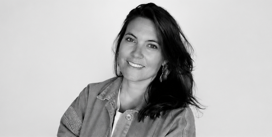 Une femme souriante avec des cheveux longs et foncés, portant un blazer en denim et des boucles d'oreilles, contre un fond blanc.