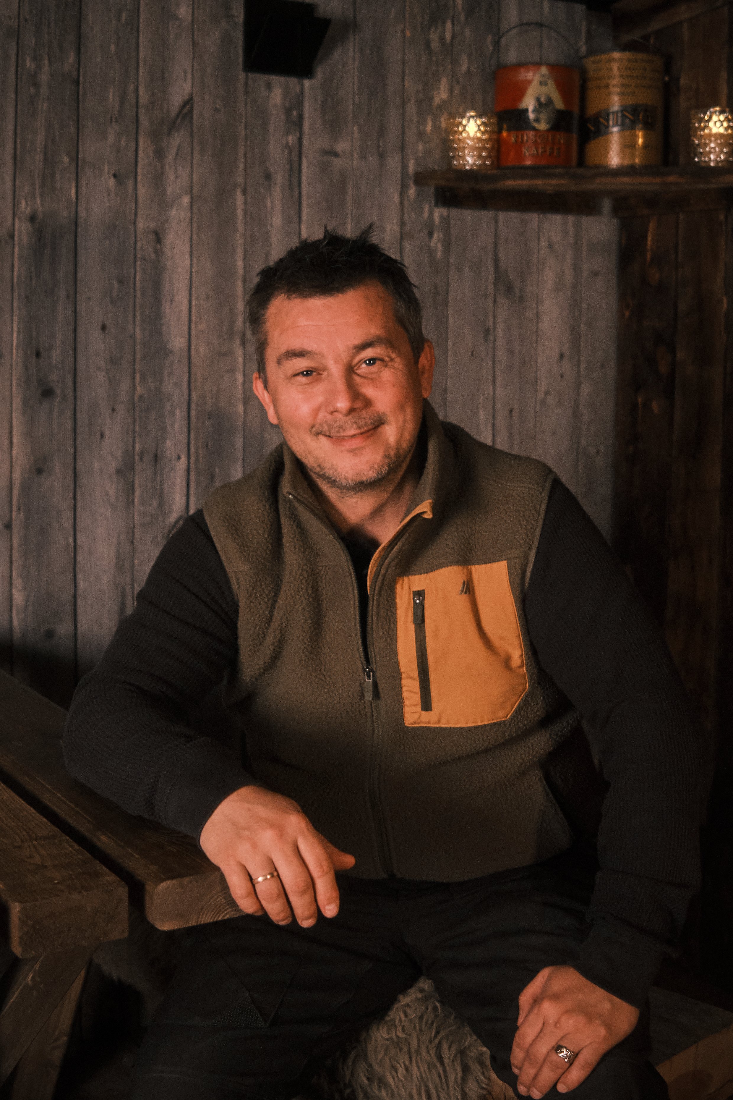 A man sitting indoors in front of a wooden wall, smiling, wearing a brown and black vest, with items on a shelf behind him.