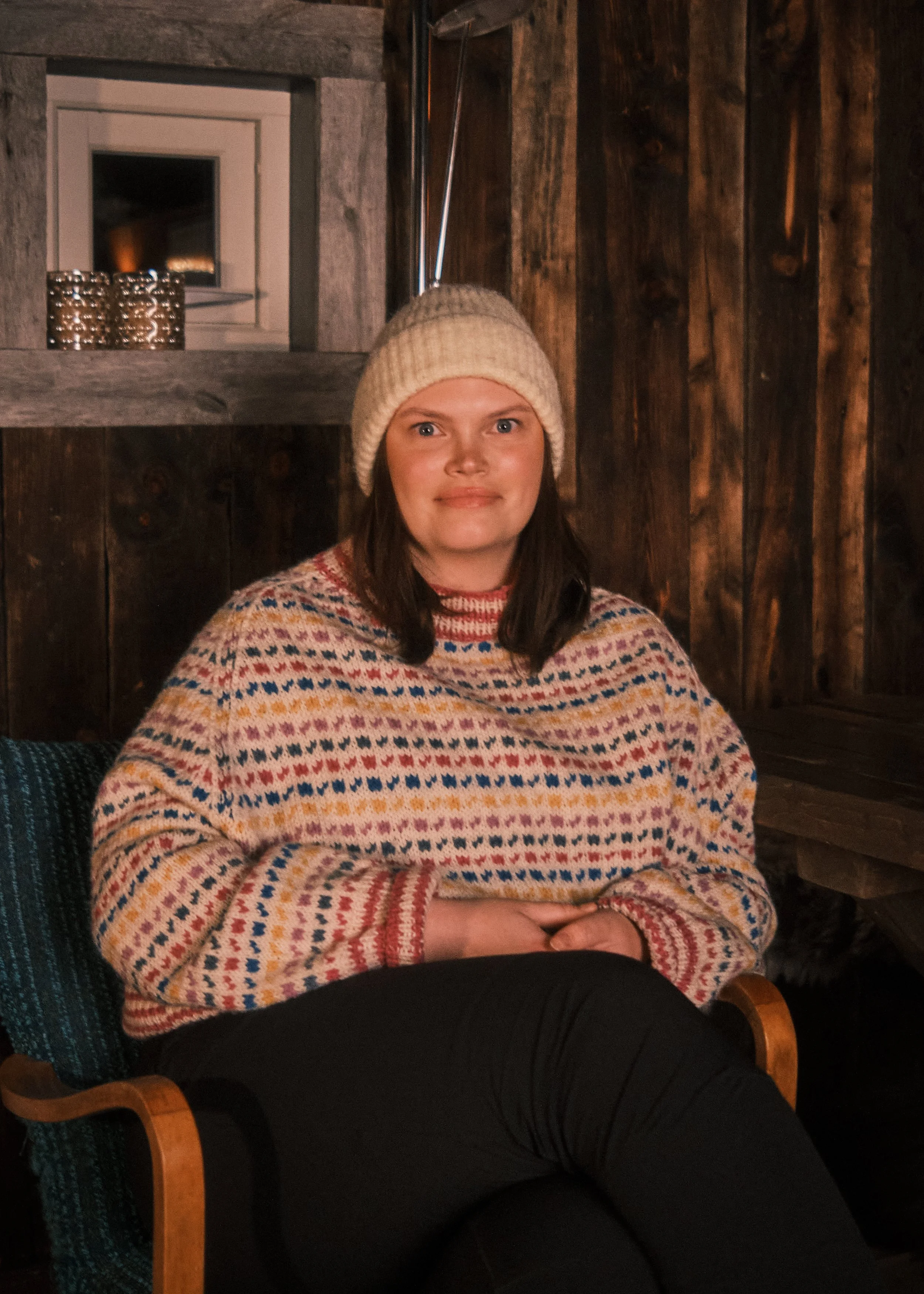 A woman wearing a knit hat and a colorful, patterned sweater sitting in a wooden room with dark wood walls and a small window behind her.