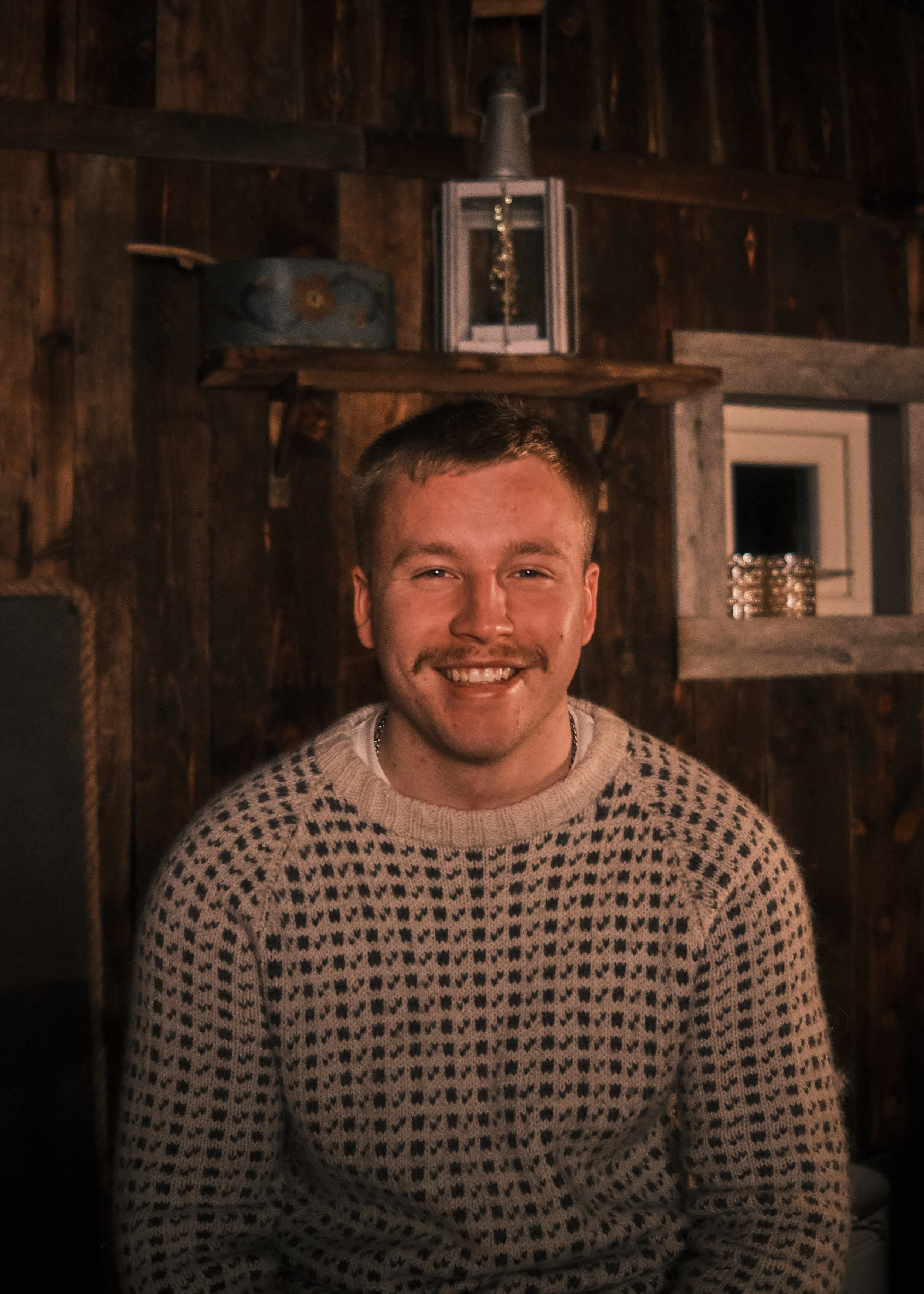 A young man smiling with a moustache, wearing a beige sweater with black hearts, sitting indoors in front of a dark wooden wall with shelves and decorations.