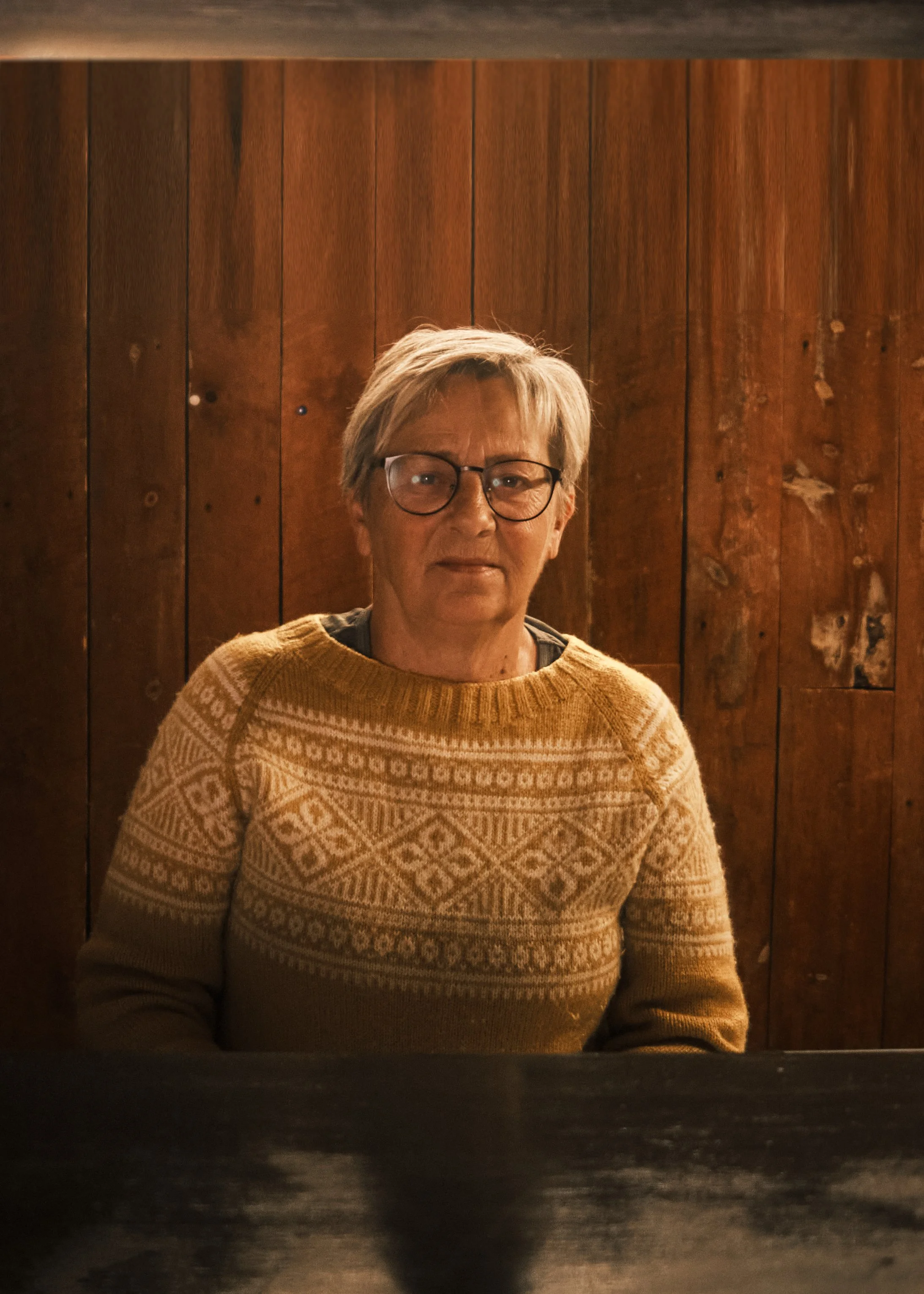 An older person with short gray hair, glasses, and a patterned beige sweater, sitting in front of a wooden wall.